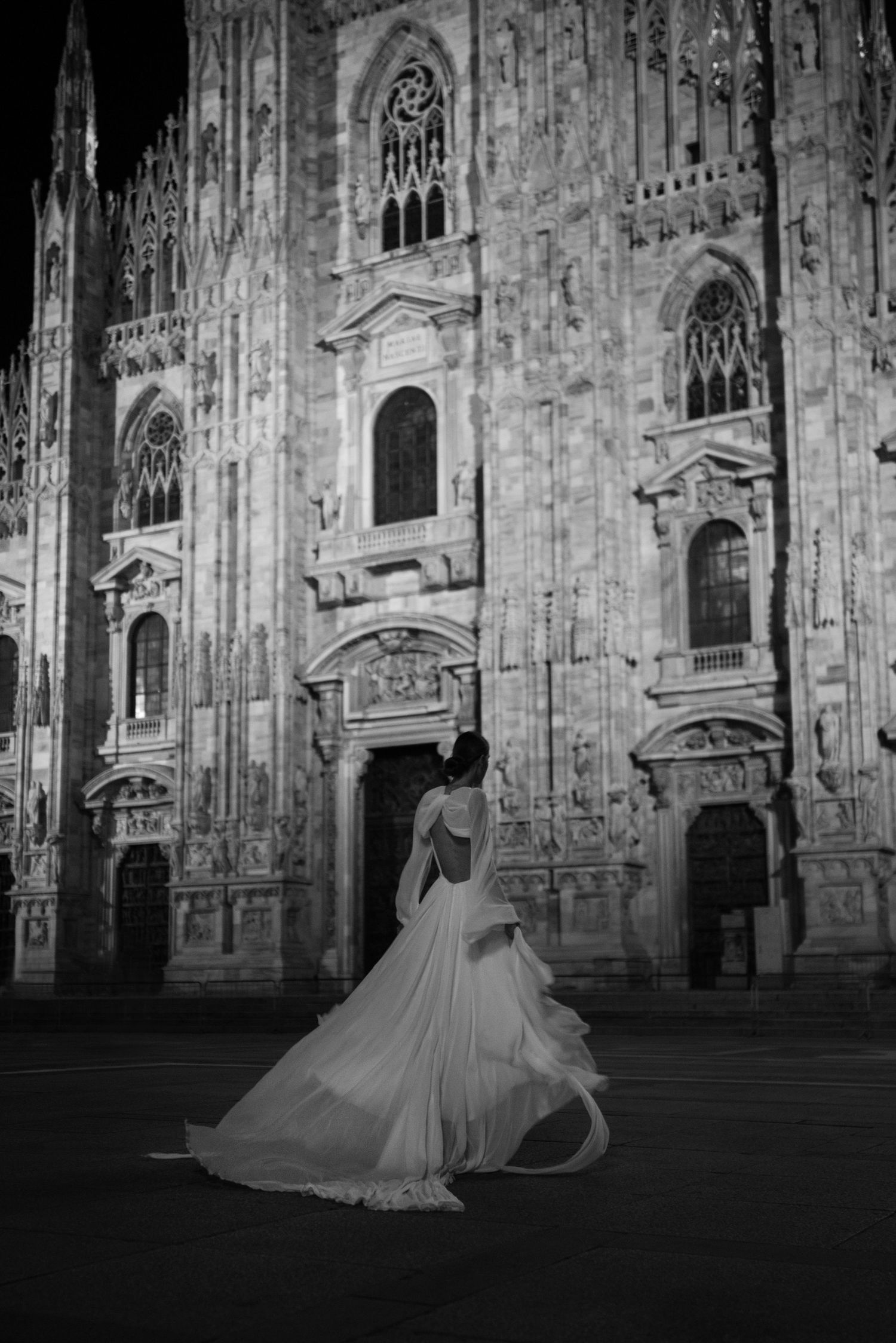 Black and white photo of a bride in flowing gown standing before Milan's illuminated Duomo Cathedral at night.