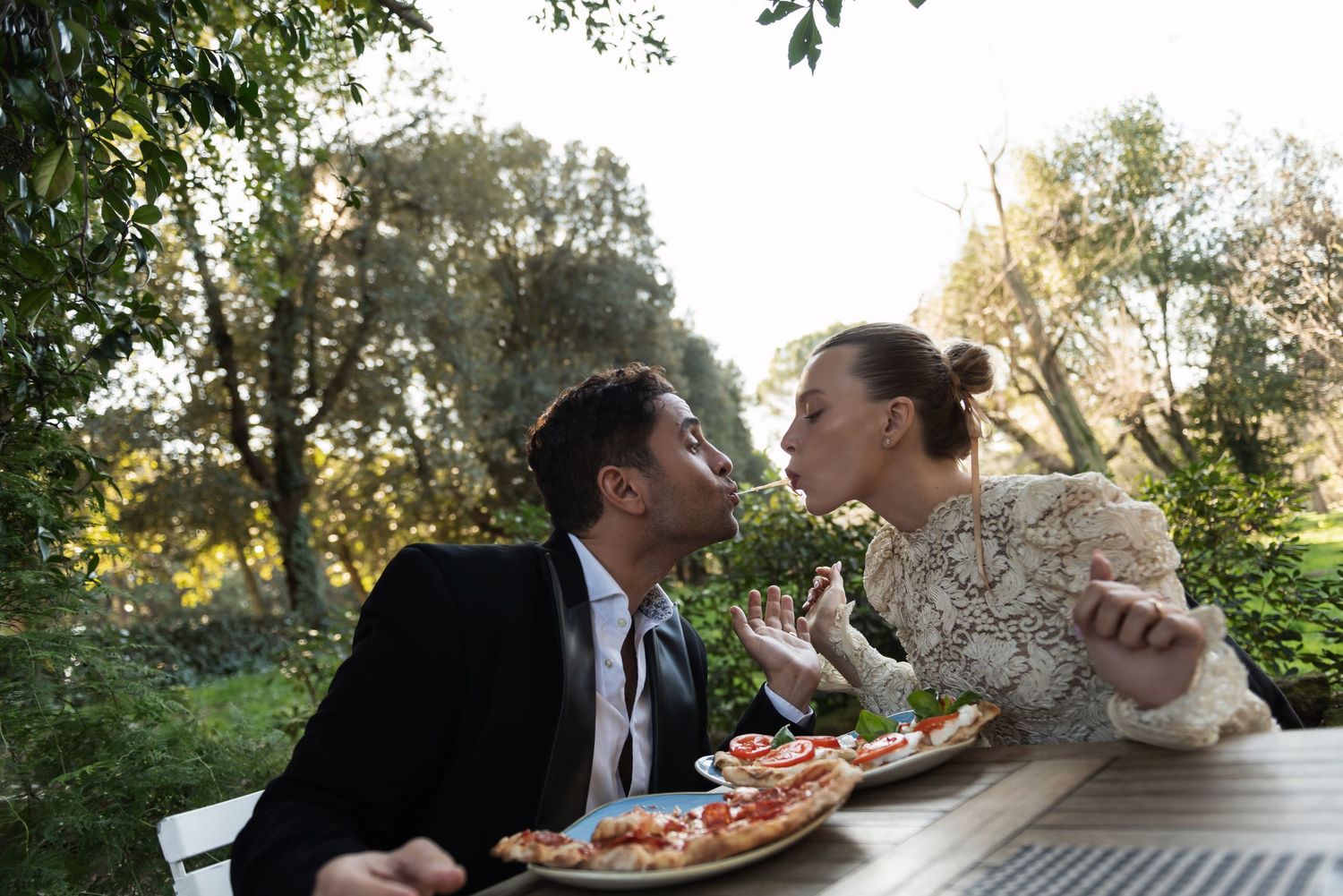 Joyful couple sharing pizza outdoors at sunlit garden table in romantic Italian setting.