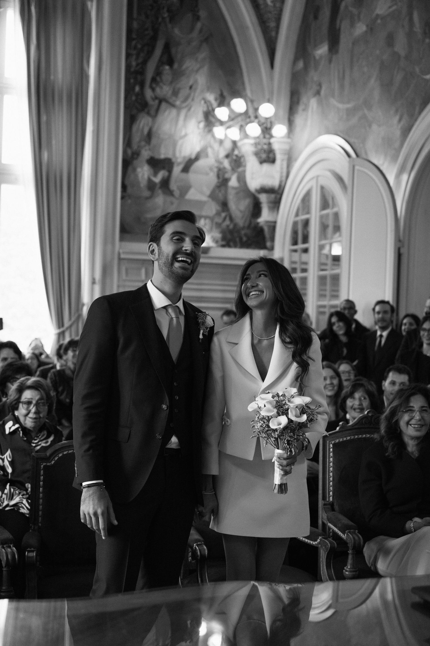 Joyful bride and groom laughing during their civil wedding ceremony in an ornate historic hall.