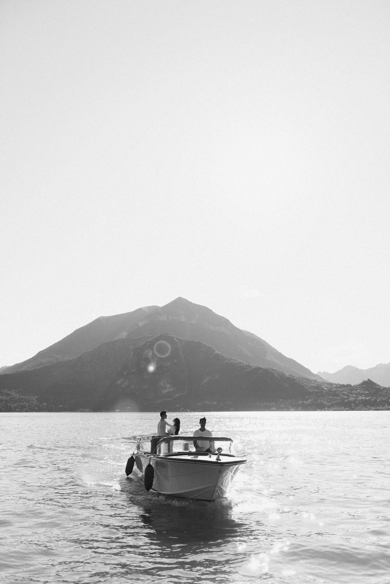 Black and white photo of couple on speedboat with mountain rising from calm lake waters. Lake Como