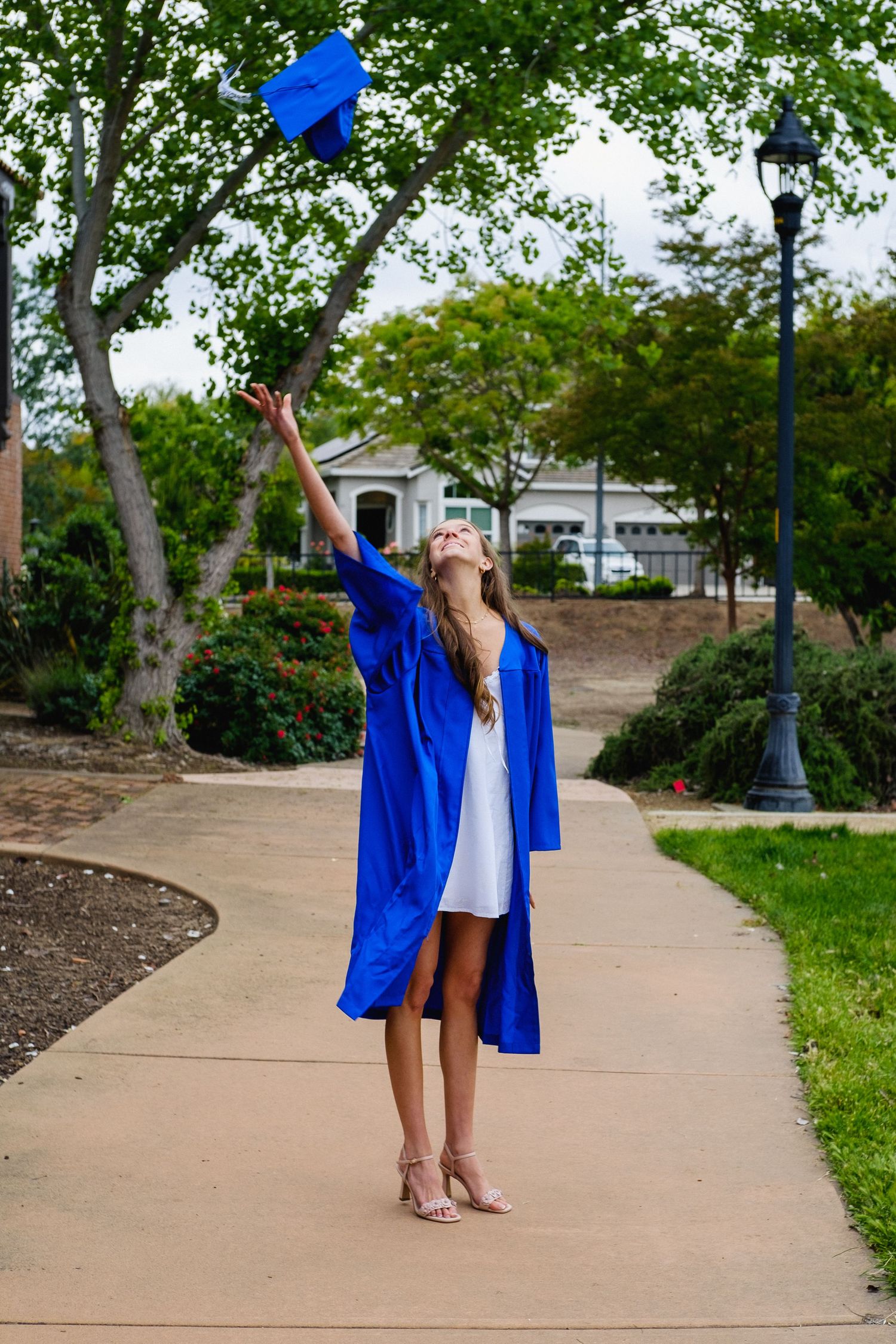 High school senior celebrating her graduation with a blue cap toss on a tree-lined path in San Jose