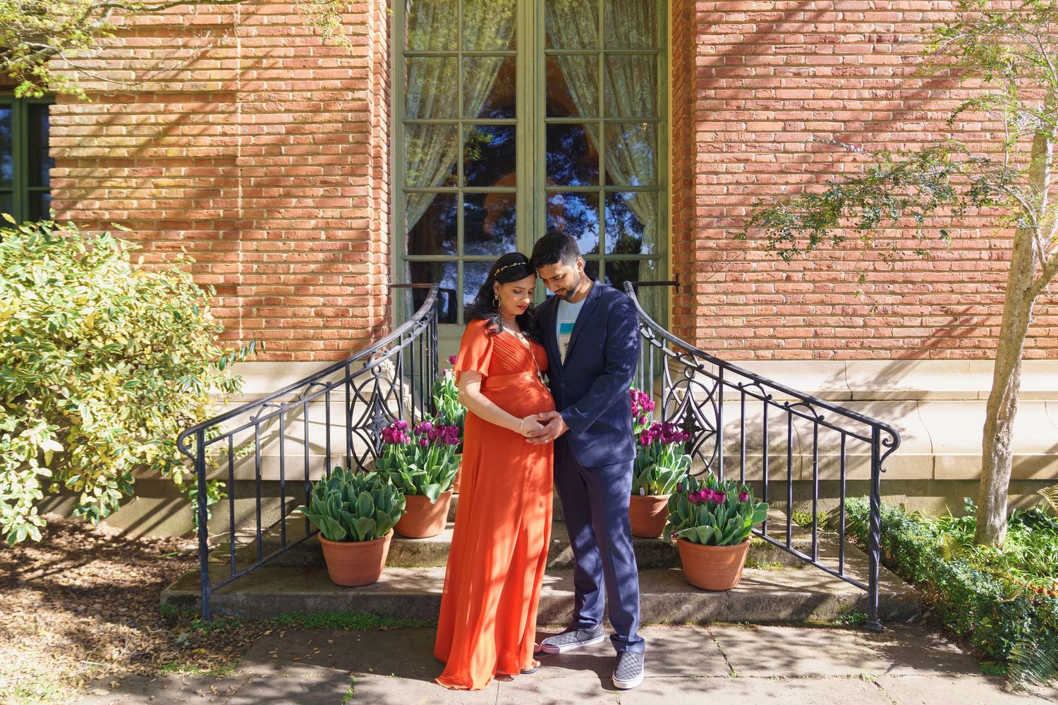 Vibrant maternity session in a Filoli garden featuring an expectant mother in a bright orange dress by an architectural window