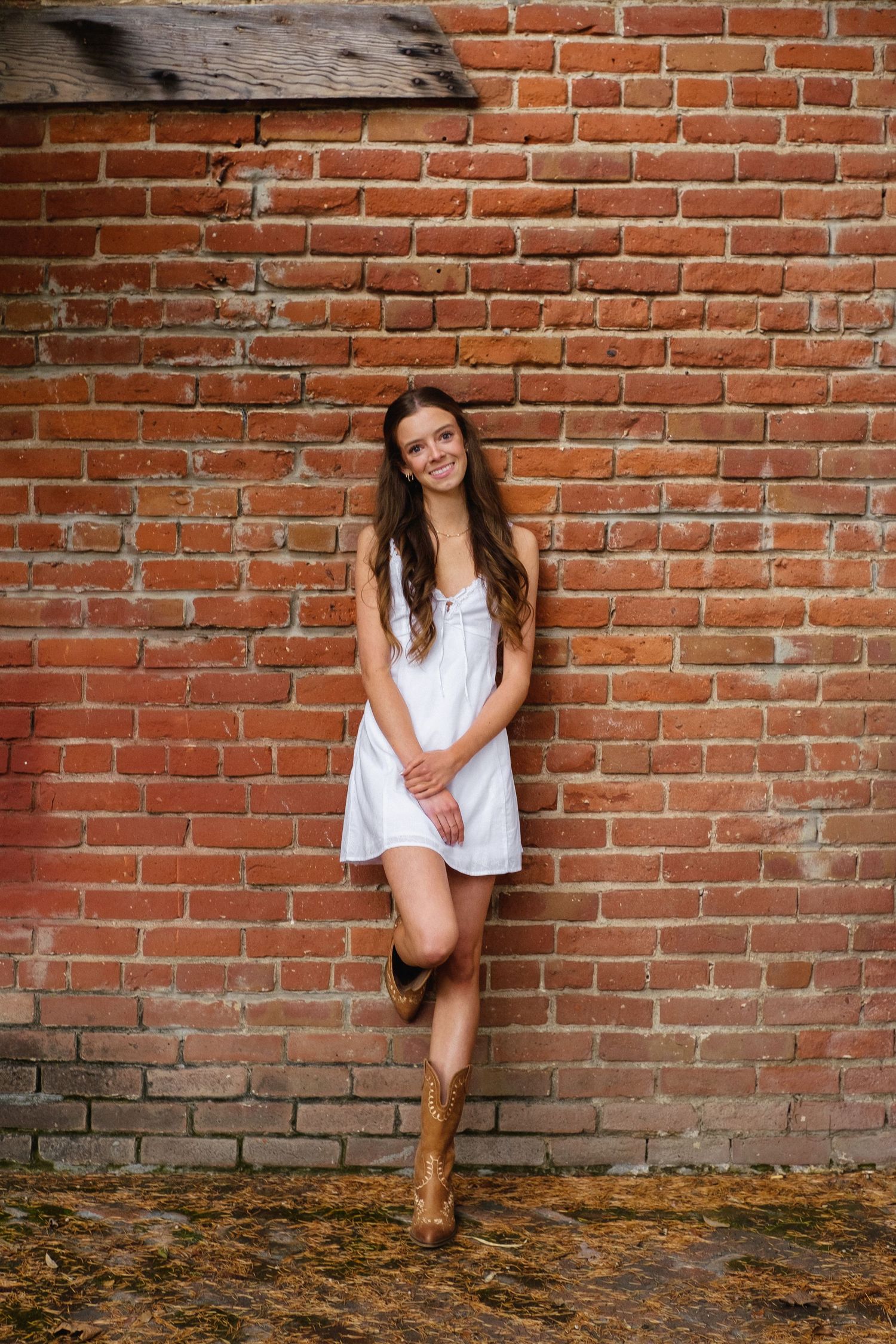 Casual senior portrait of a graduate in a white dress leaning against a rustic brick wall in San Jose