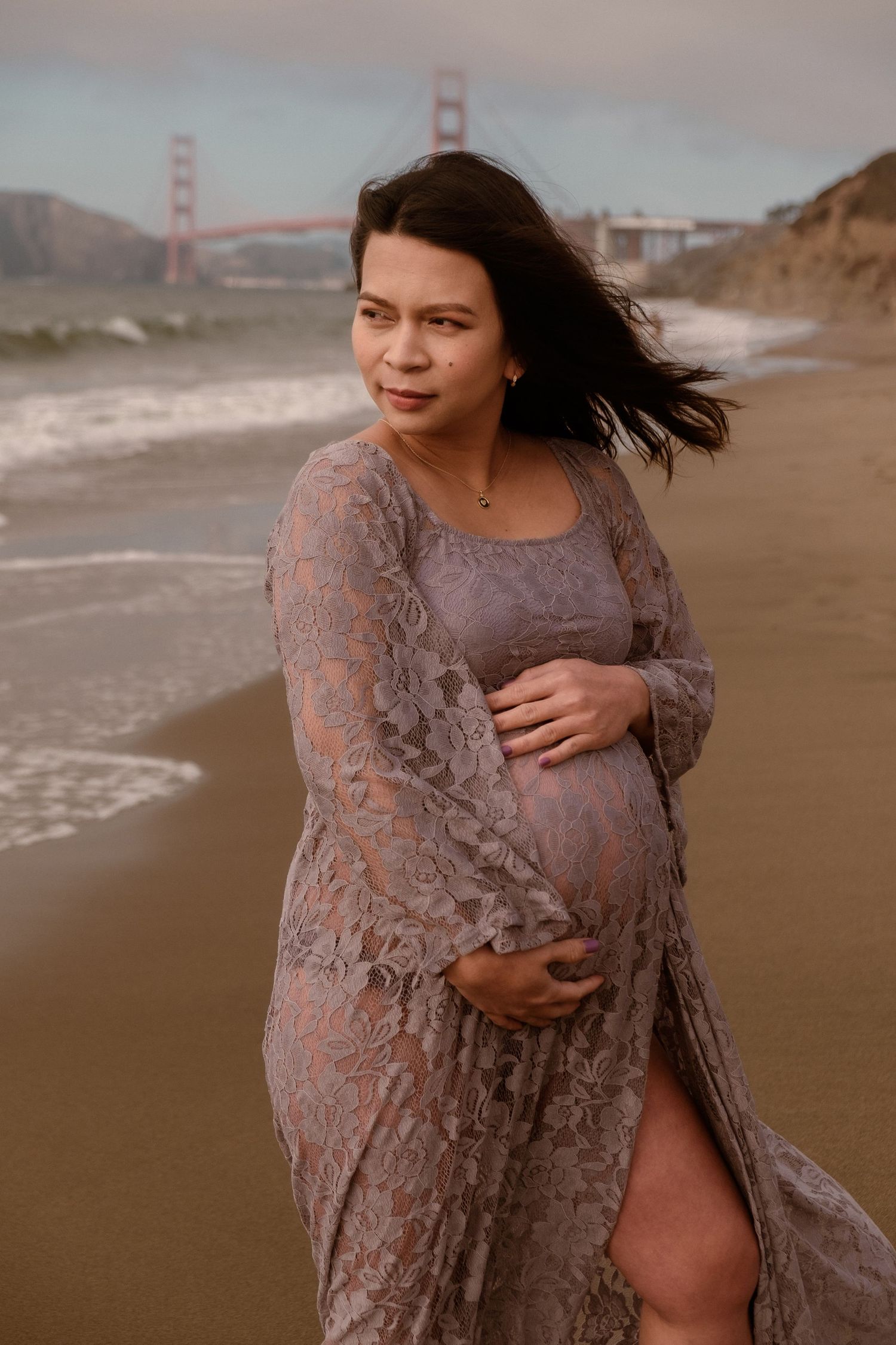 Editorial maternity portrait of a woman in a lace lavender gown on a foggy Baker Beach with golden gate bridge in the background