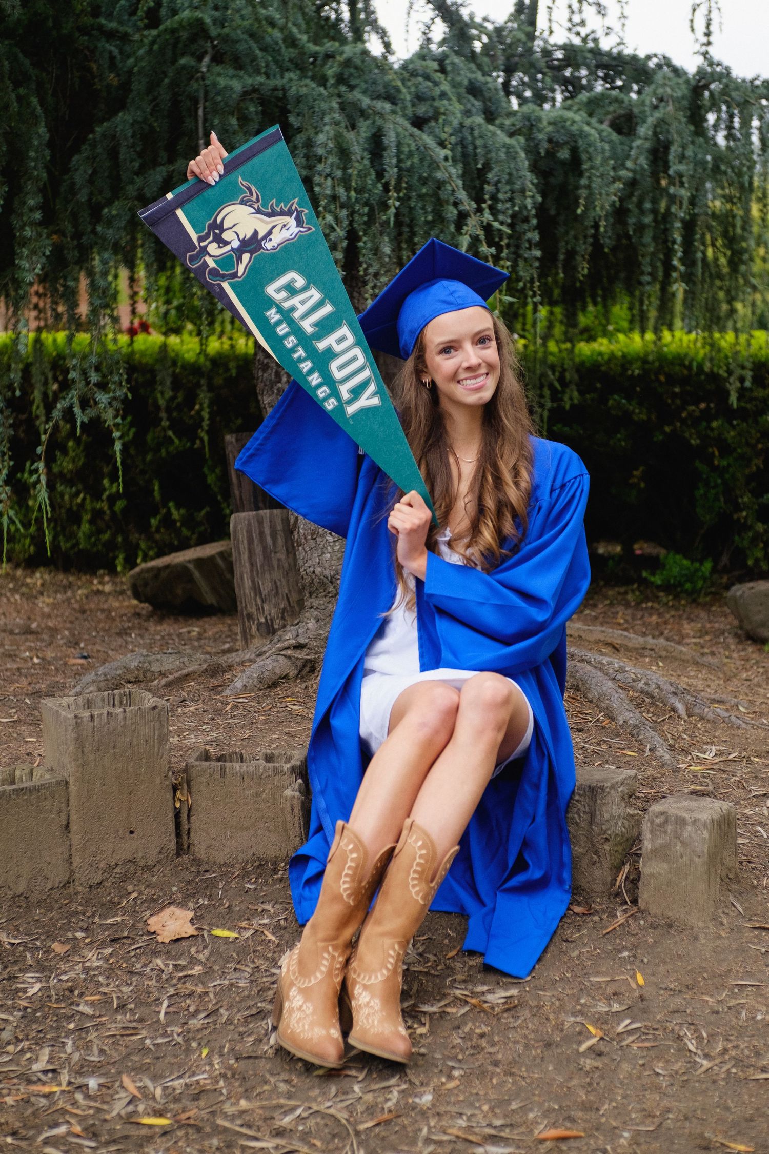 Senior graduation portrait featuring a student in a blue gown holding a college pennant and wearing cowboy boots