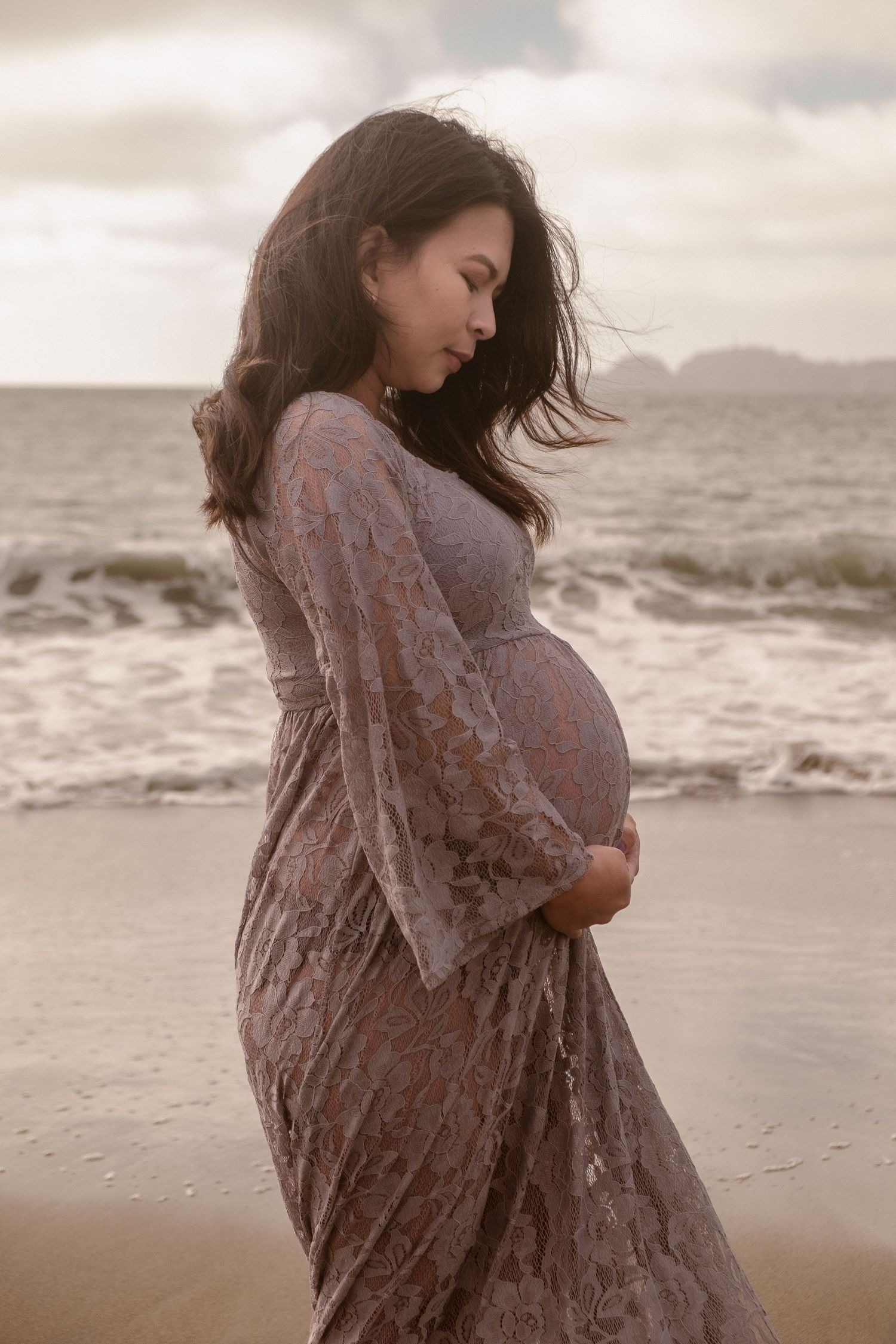 Editorial maternity portrait of a woman in a lace lavender gown on a foggy Bay Area beach