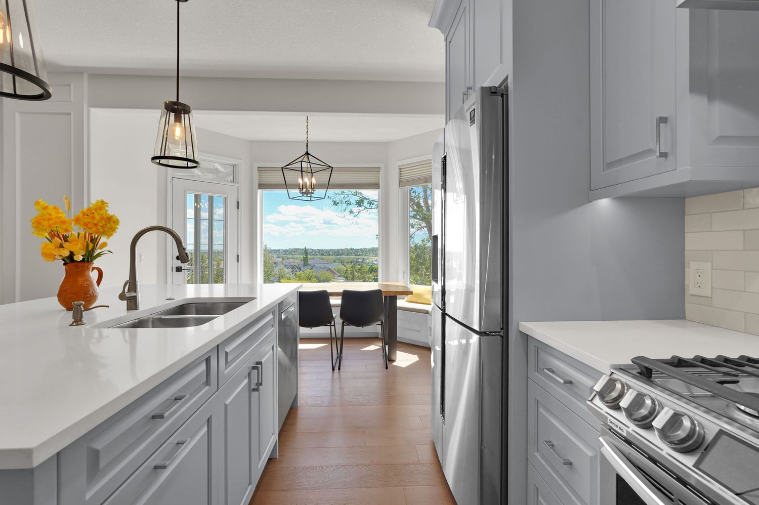 Bright white kitchen with gray cabinets, quartz countertops, gas range, and scenic views through large windows.