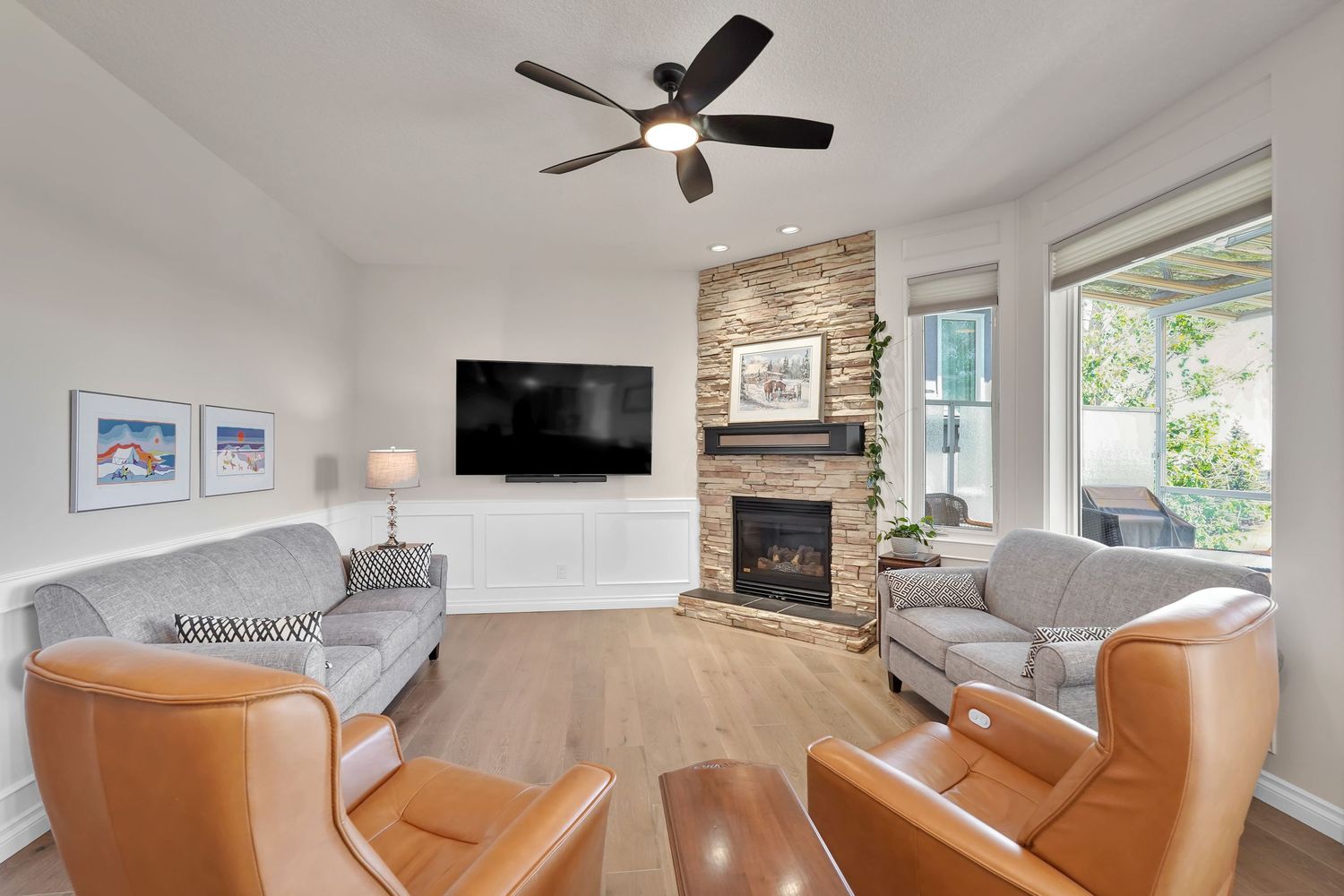 Cozy living room with stone fireplace, wall-mounted TV, ceiling fan, hardwood floors, and leather accent chairs.