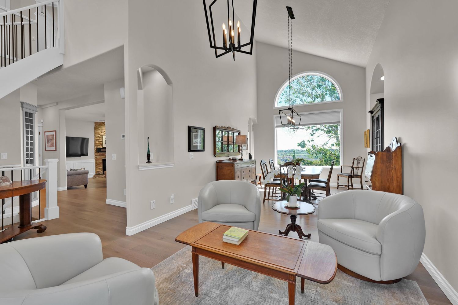 Bright living room with high ceilings, gray armchairs, wood coffee table, chandelier, and open view to dining area and large window.