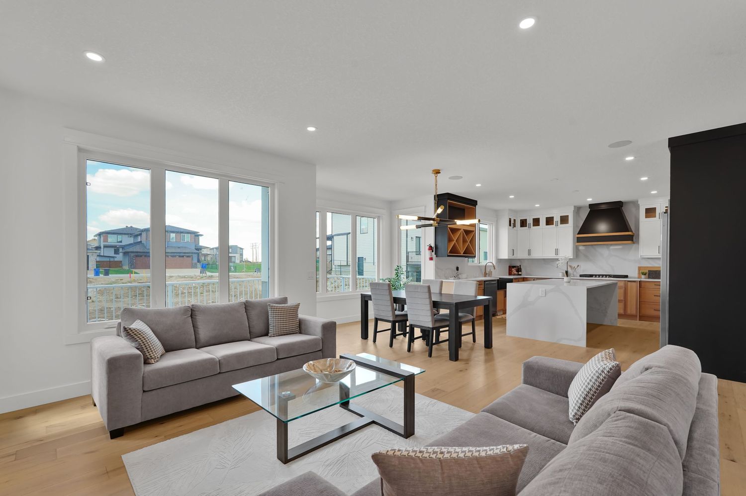 Open-concept living and dining area with gray sofas, kitchen island, and large windows with natural light.
