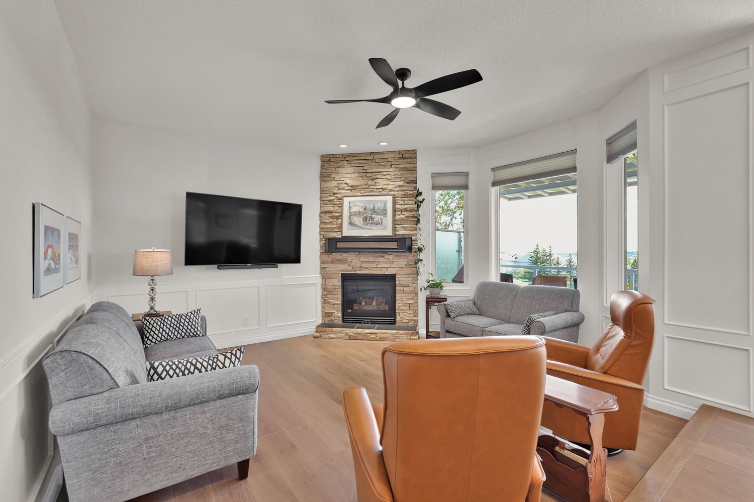 Modern living room with stone fireplace, ceiling fan, gray sofas, leather chairs, and hardwood floors.