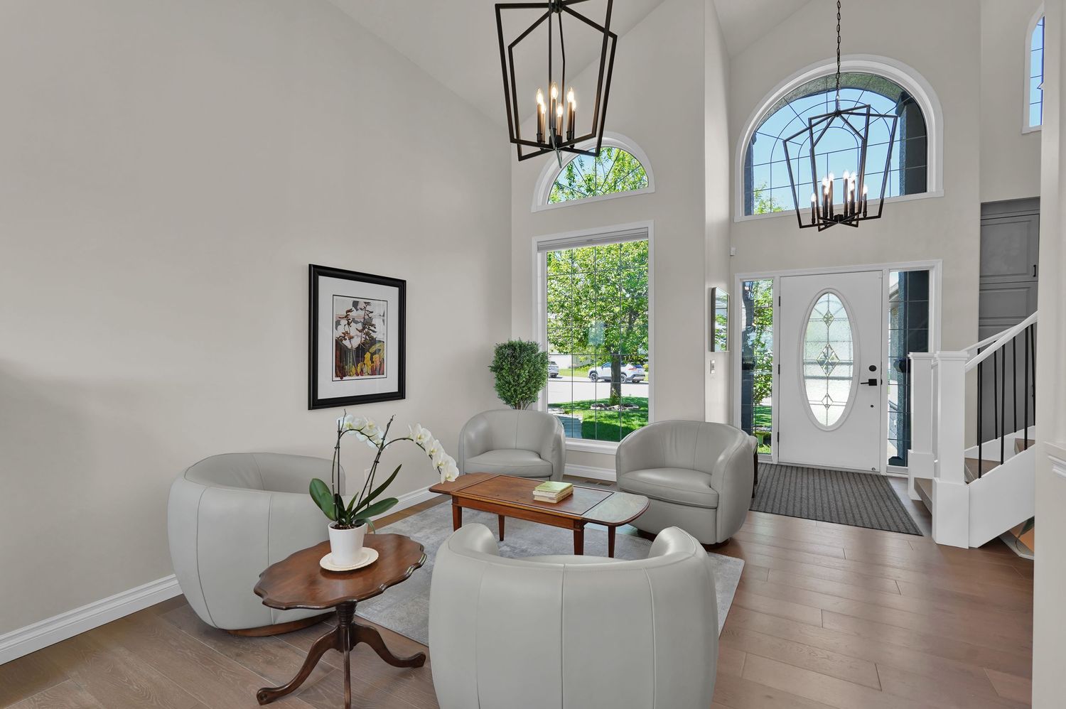 Bright two-story living room with vaulted ceilings, chandelier, gray chairs, hardwood floors, and arched front door.