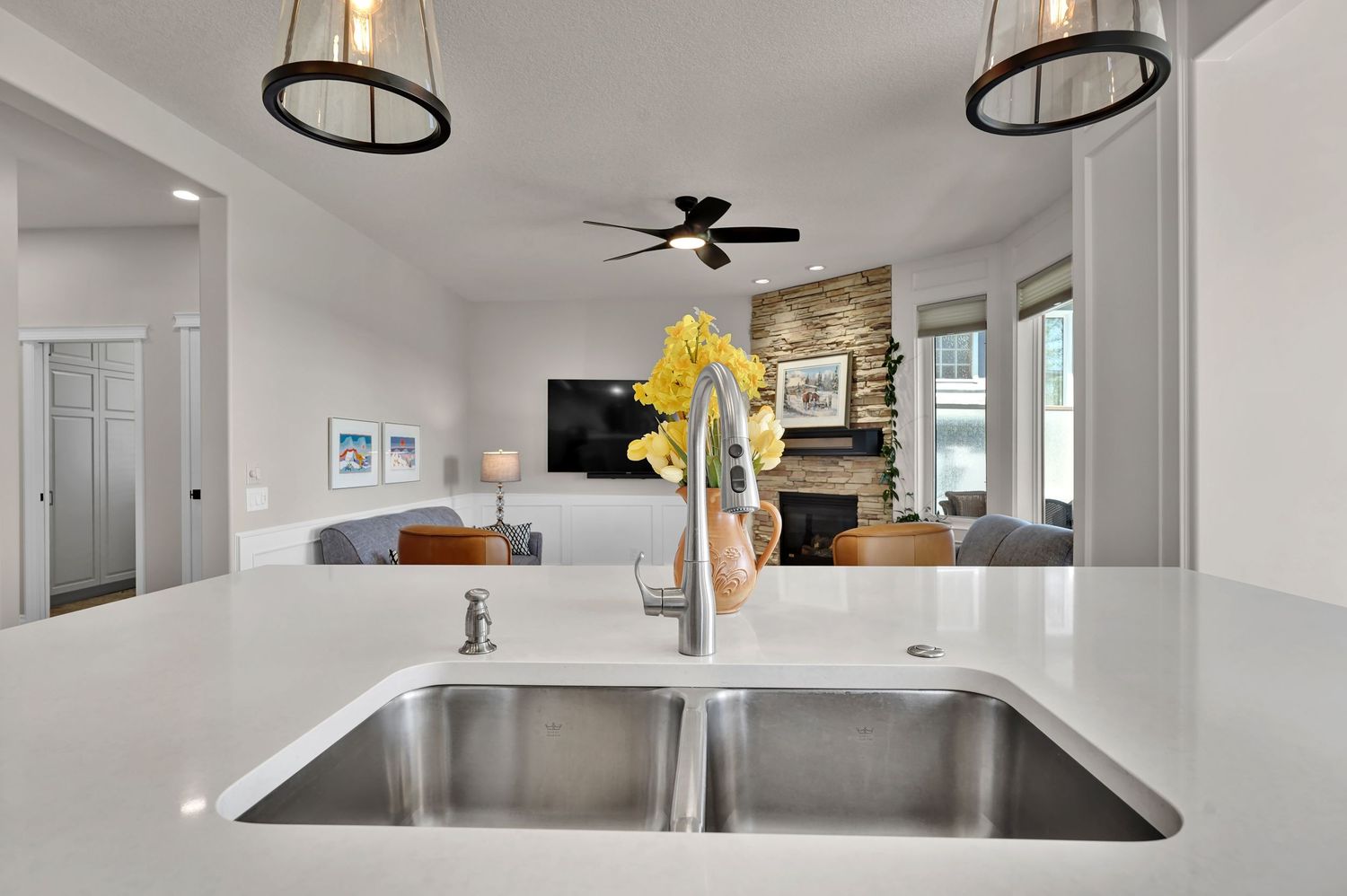 Modern kitchen with stainless steel double sink, white quartz countertop, open to living room with stone fireplace.