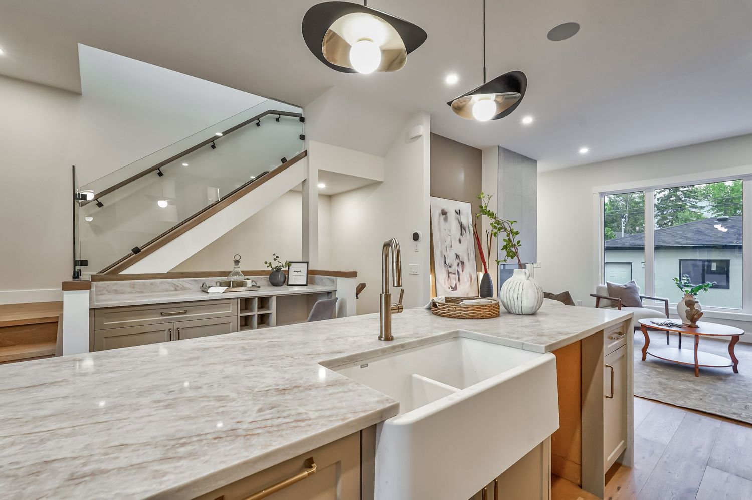 Modern kitchen with marble island, farmhouse sink, pendant lights, and open floor plan with staircase in background.
