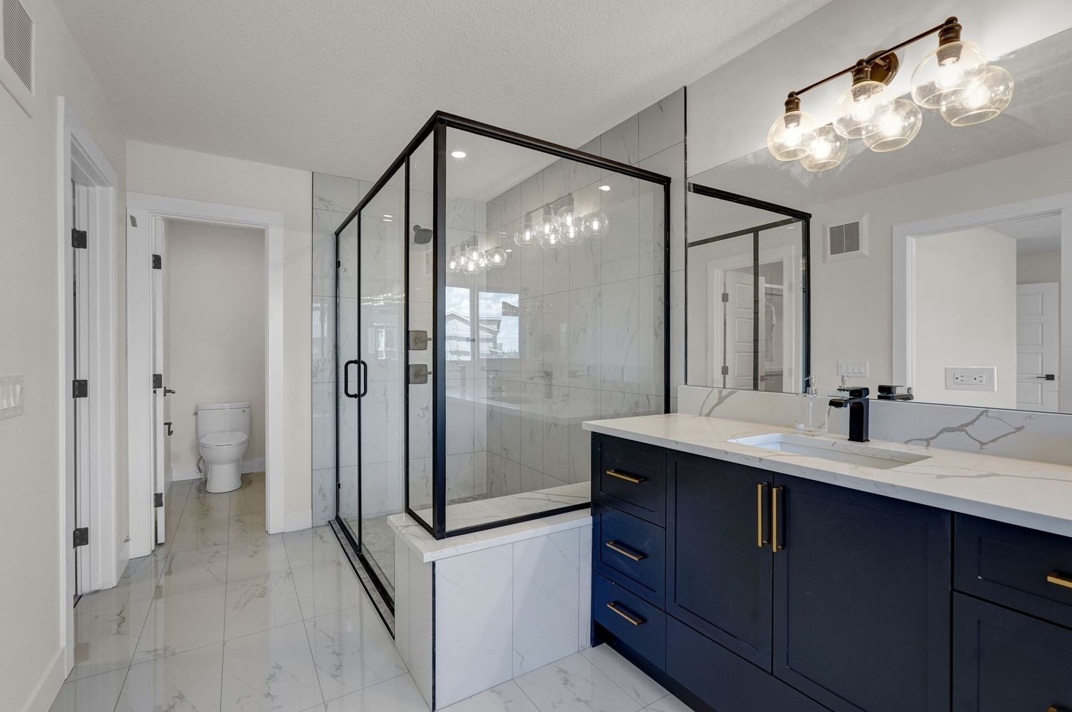 Modern bathroom with navy blue vanity, gold hardware, glass shower enclosure, and marble tile flooring.