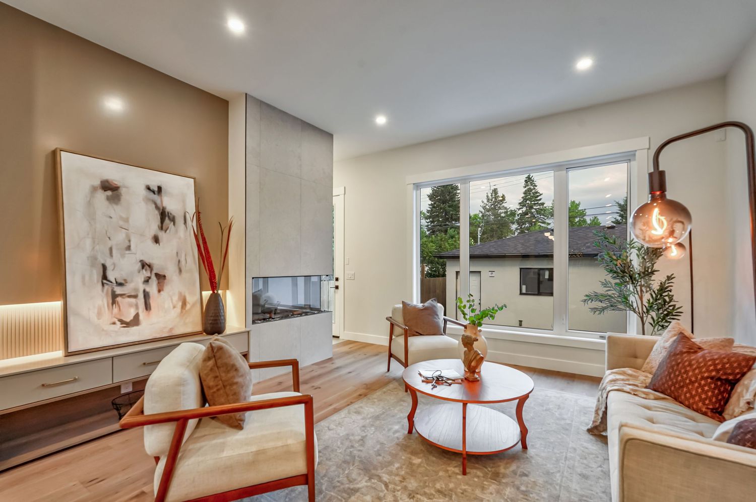 Modern living room with white chairs, oval coffee table, large window, wall art, and copper accent lamp on hardwood floors.