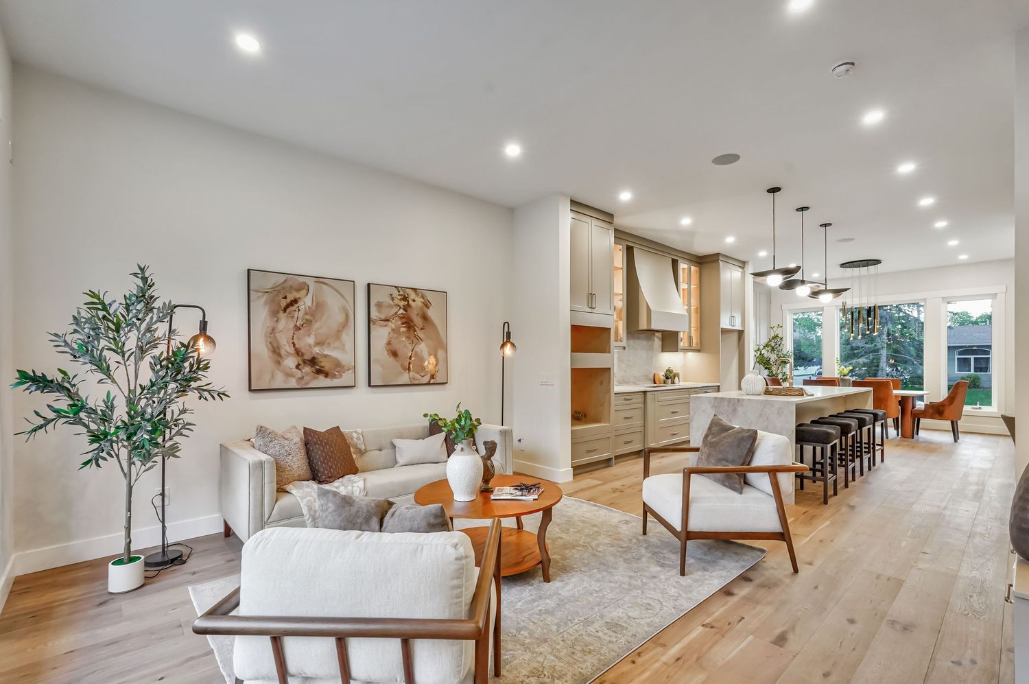 Modern open-concept living room with light wood floors, neutral tones, mid-century furniture, and a bright kitchen in the background.