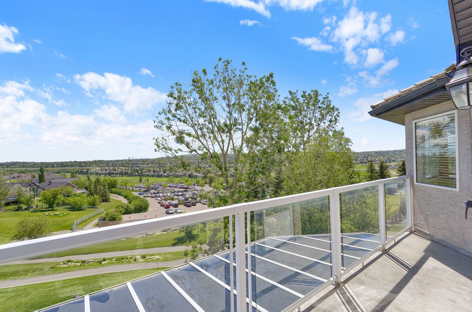 A spacious balcony with glass railings offers panoramic views of a lush green neighborhood under a bright blue sky.