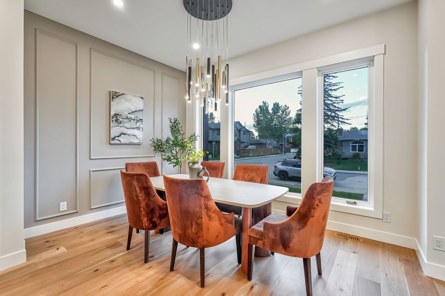 Modern dining room with russet velvet chairs, white table, pendant lighting, and large window overlooking trees.