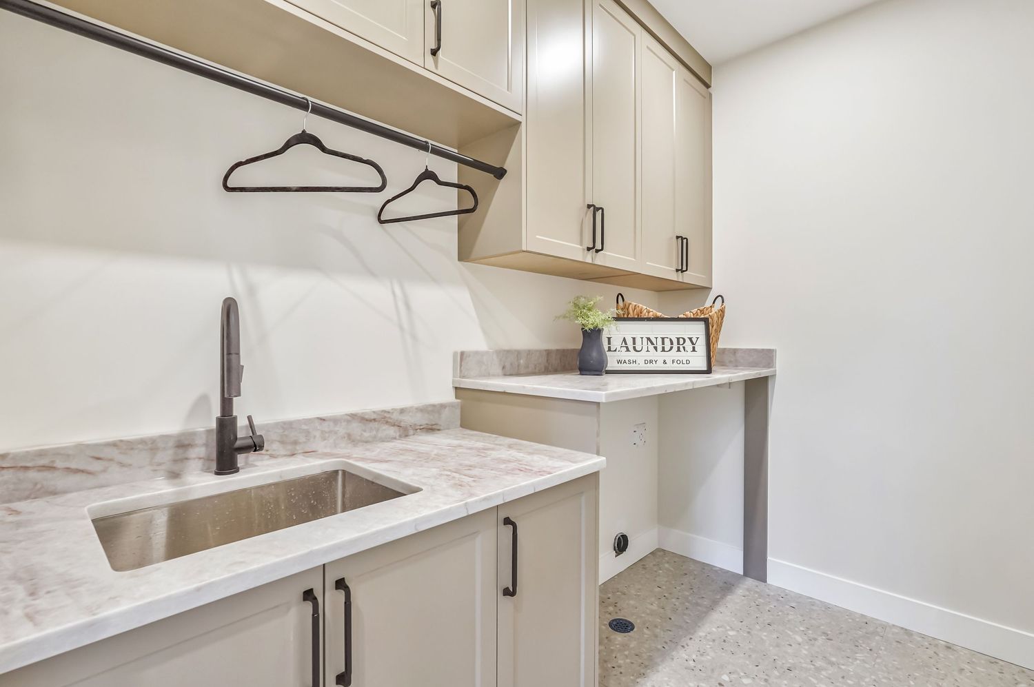 Modern laundry room with marble countertop, undermount sink, beige cabinets, hanging rod, and built-in desk area.