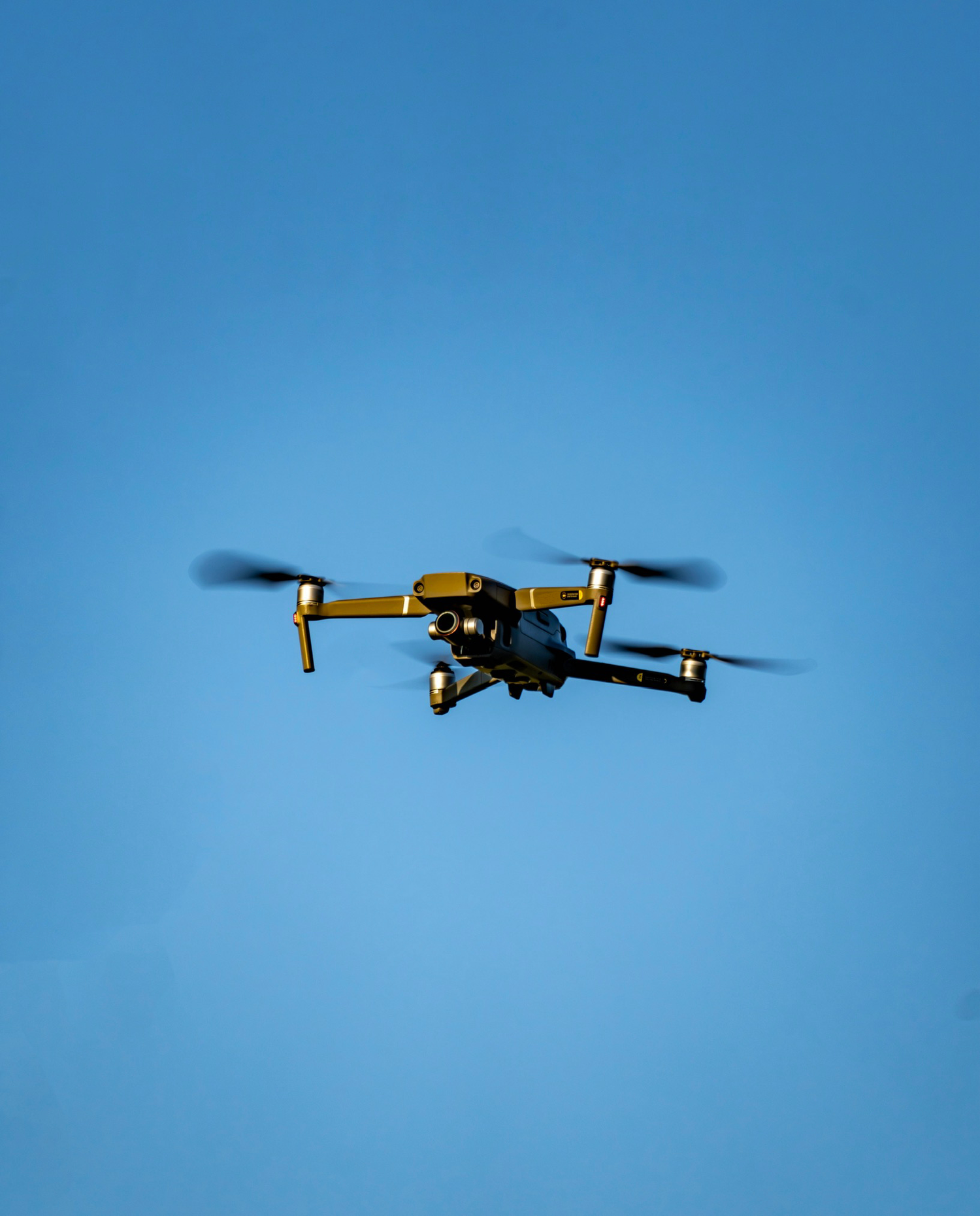 Gold and black quadcopter drone captured mid-flight against a vivid clear blue sky.