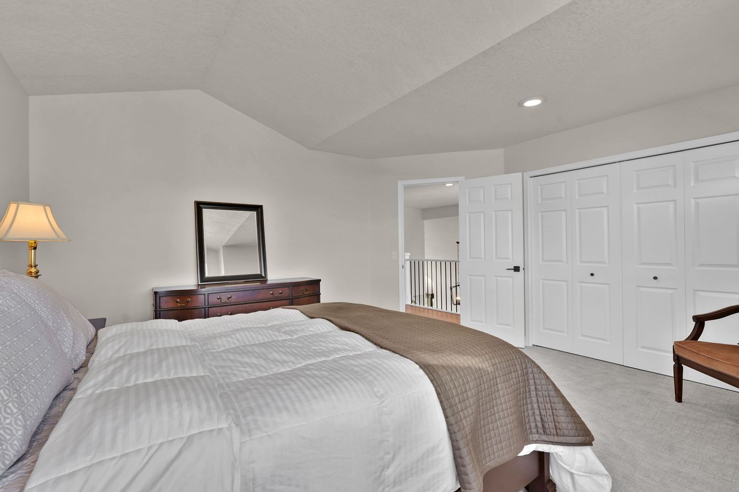 Spacious master bedroom with vaulted ceiling, plush carpet, dark wood headboard, white closet doors, and soft lamp lighting.