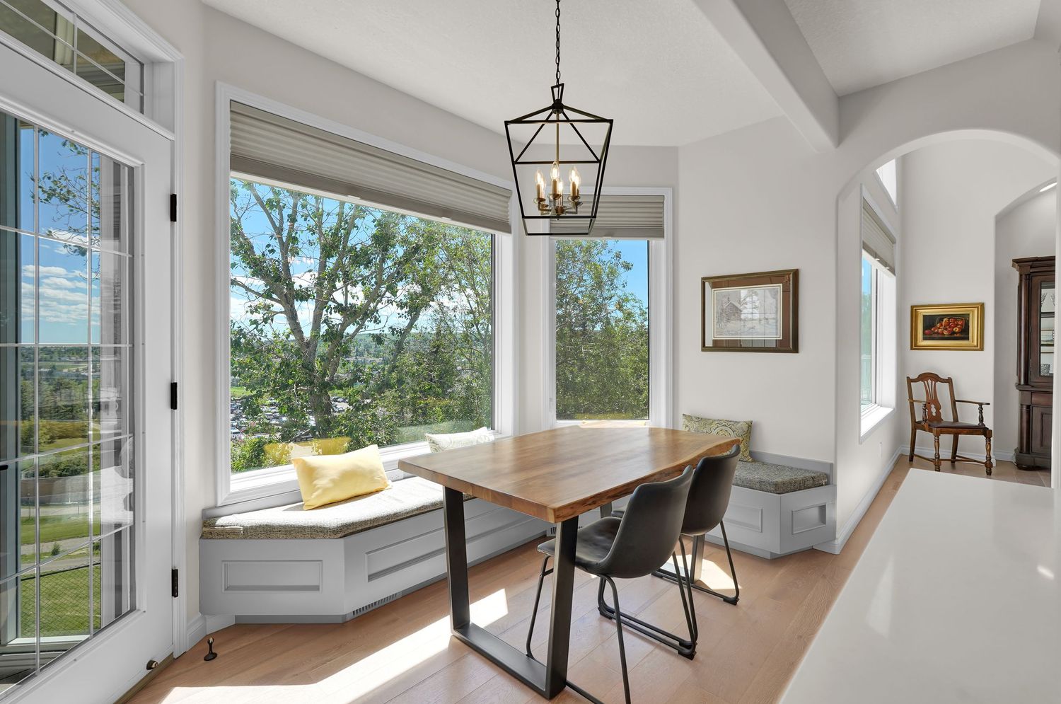 Bright dining nook with built-in window seat, wooden table, modern chairs, and a chandelier overlooking lush green trees.