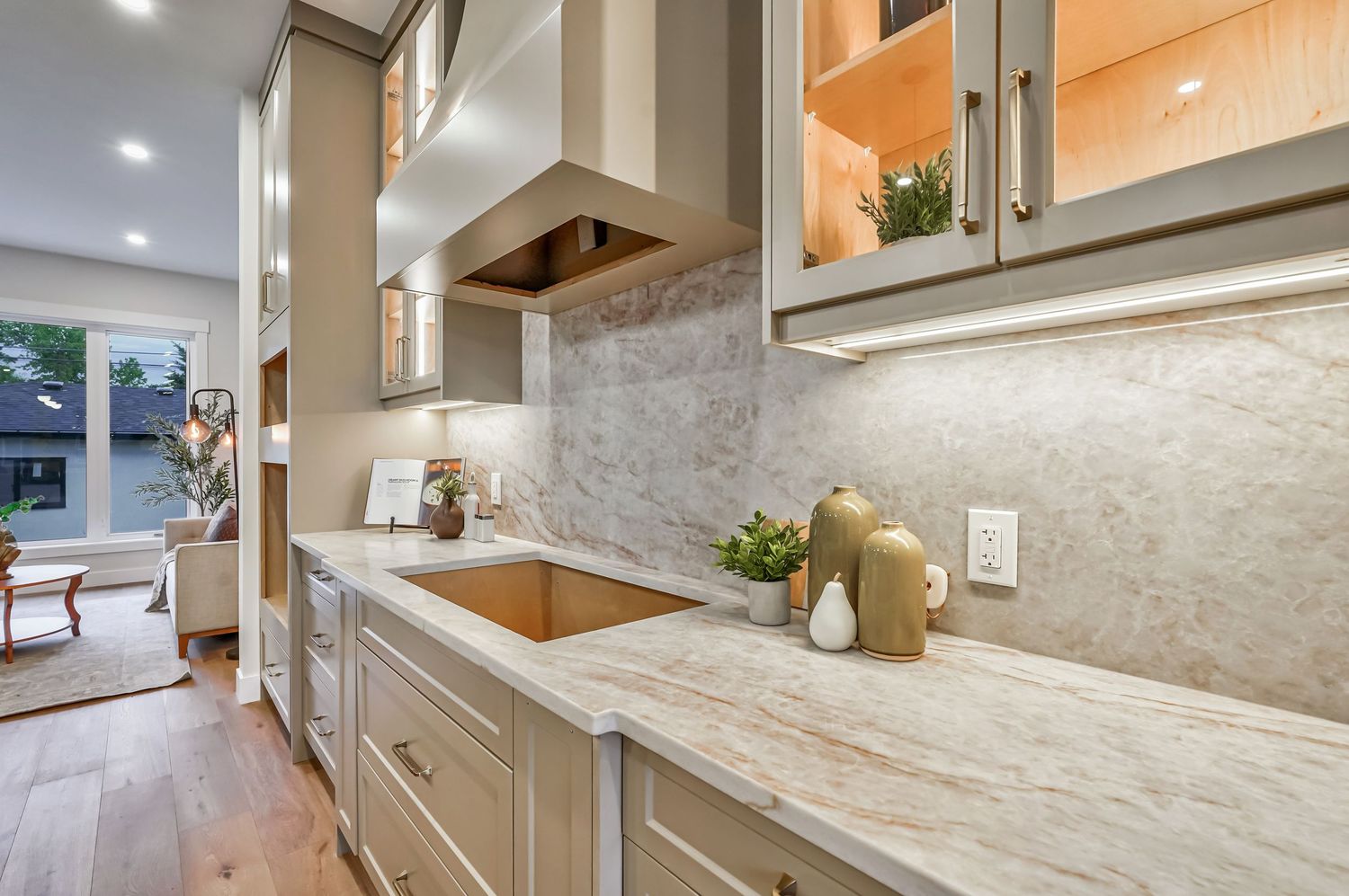 Elegant kitchen featuring marble backsplash, quartz countertops, taupe cabinetry, and a copper sink with under-cabinet lighting.
