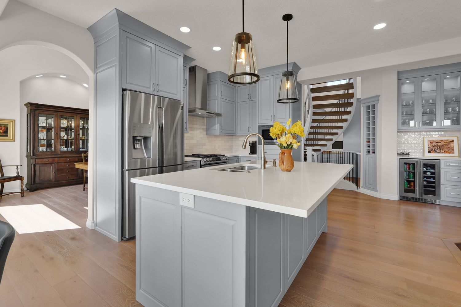 Modern kitchen with gray cabinets, white island, pendant lights, stainless steel fridge, and hardwood floors.