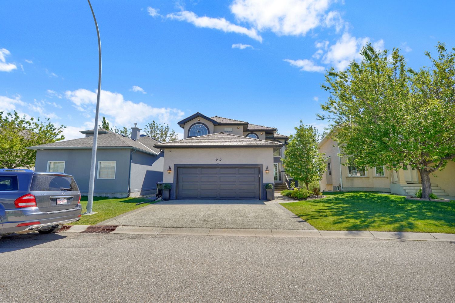 Two-story suburban home with gray garage door, green lawn, and mature trees under a bright blue sky.