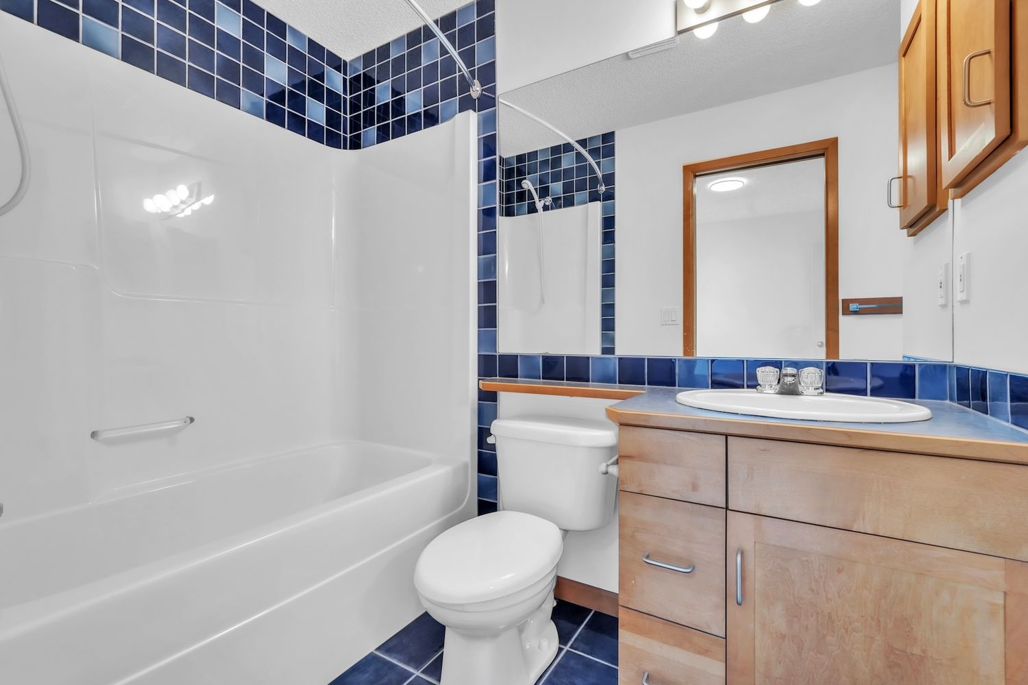 Full bathroom with blue tile accents, white tub-shower combo, toilet, and wood vanity with mirror.