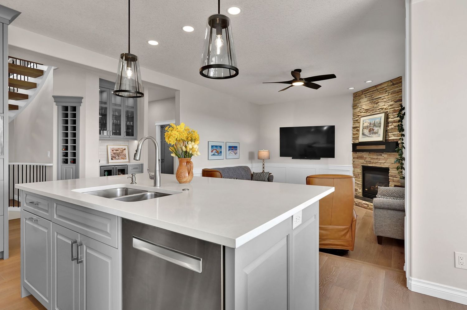 Modern kitchen island with white quartz countertop, gray cabinets, and pendant lights opening to a cozy living room with stone fireplace.