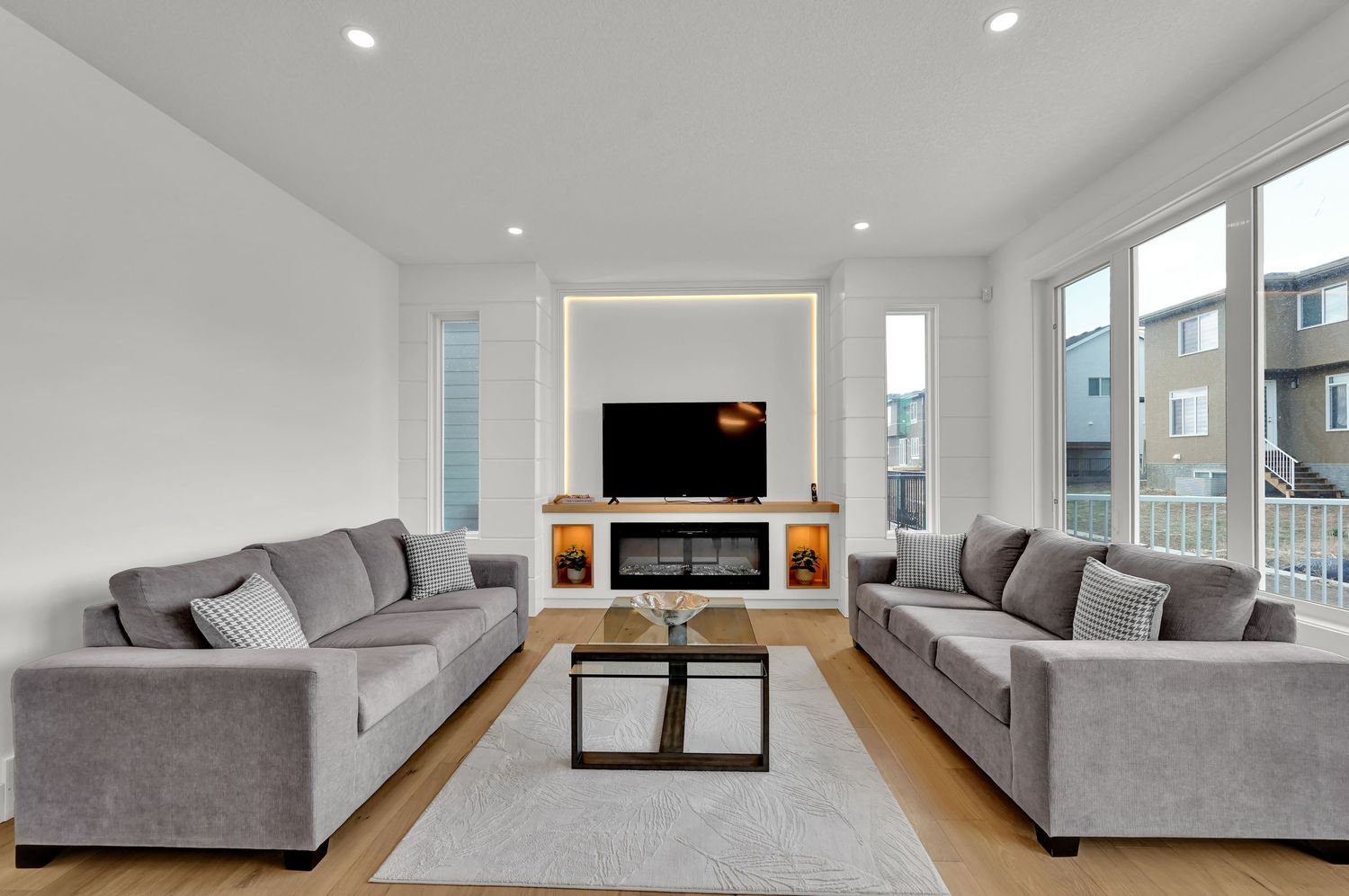 Modern living room with gray sofas, glass coffee table, electric fireplace, and wall-mounted TV on light hardwood floors.