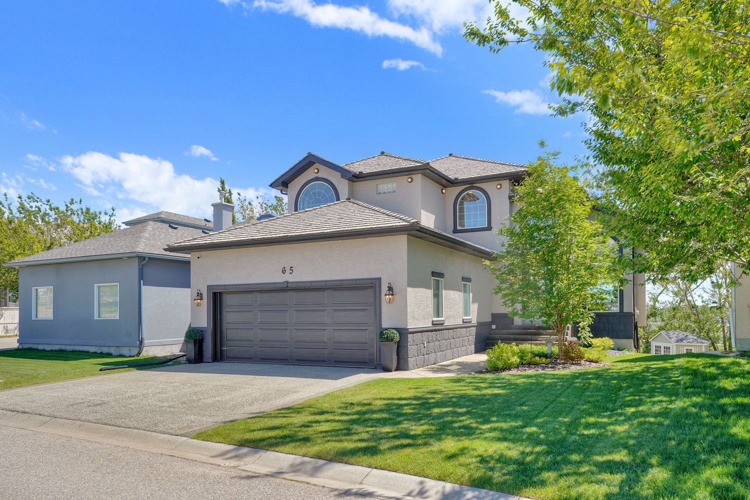 Two-story beige stucco home with brown garage door, arched windows, and lush green trees on a sunny day.