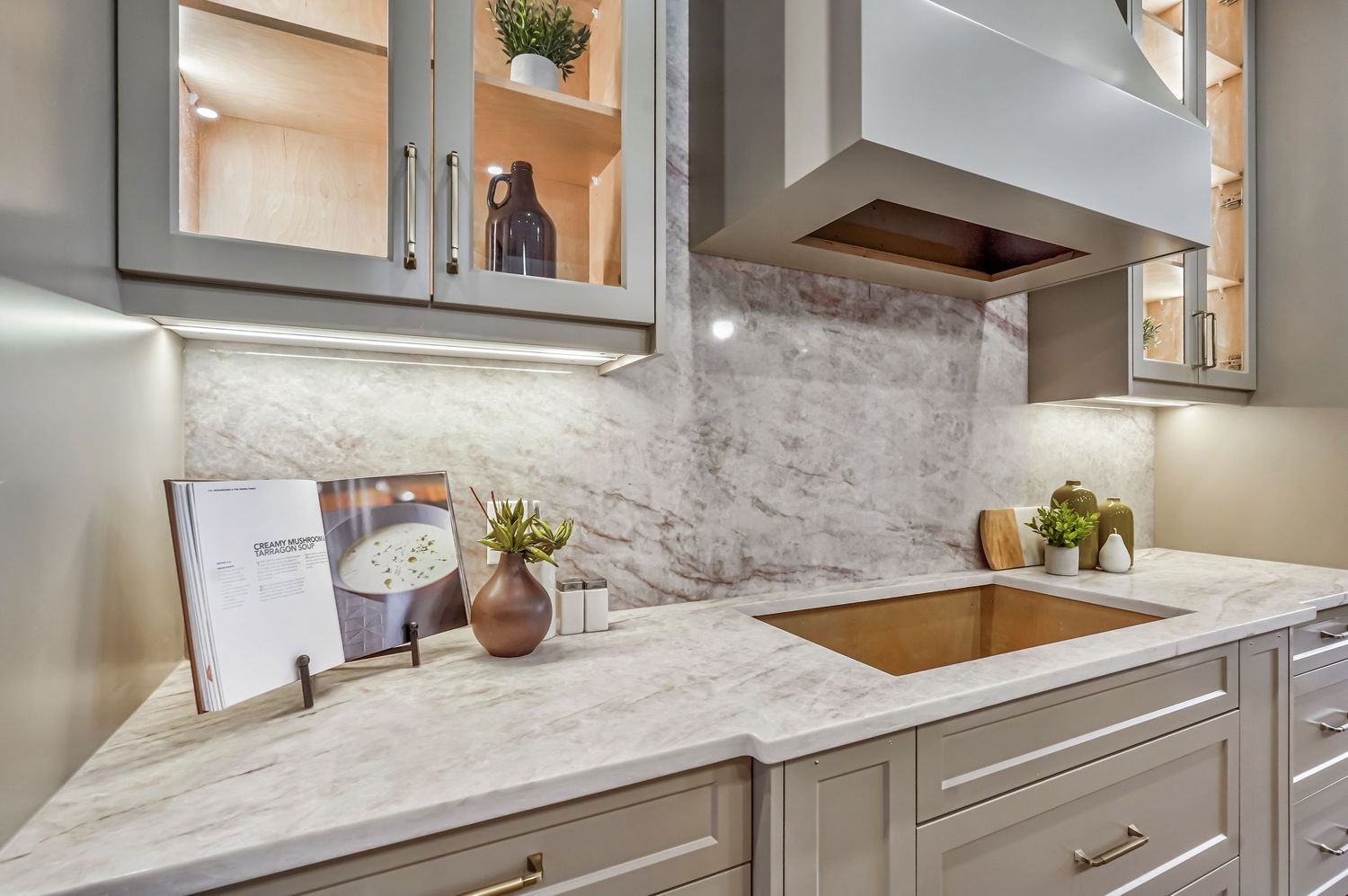 Modern kitchen with marble countertops, gray cabinets, gold hardware, and a sleek range hood over a copper sink.