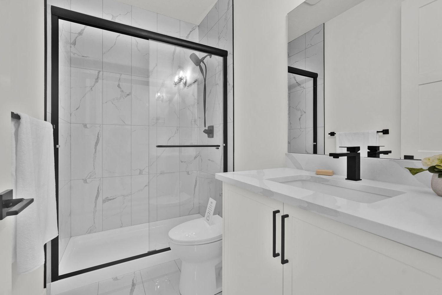 Clean white bathroom with marble-look tiled shower, black-framed glass door, and single vanity with black fixtures.