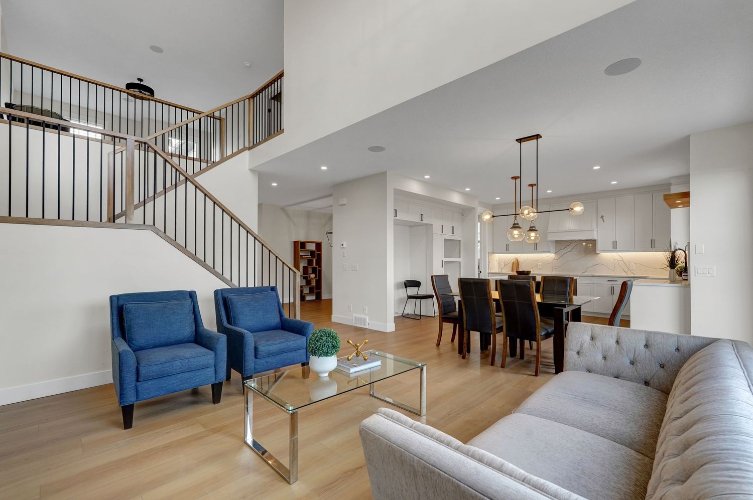 Open-concept living space with gray sofa, blue chairs, staircase, and view into modern white kitchen.