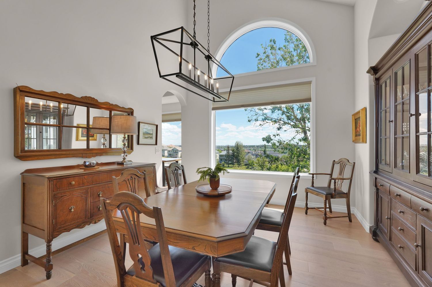 Elegant dining room with wooden furniture, large arched window, scenic view, and modern chandelier on light hardwood floors.