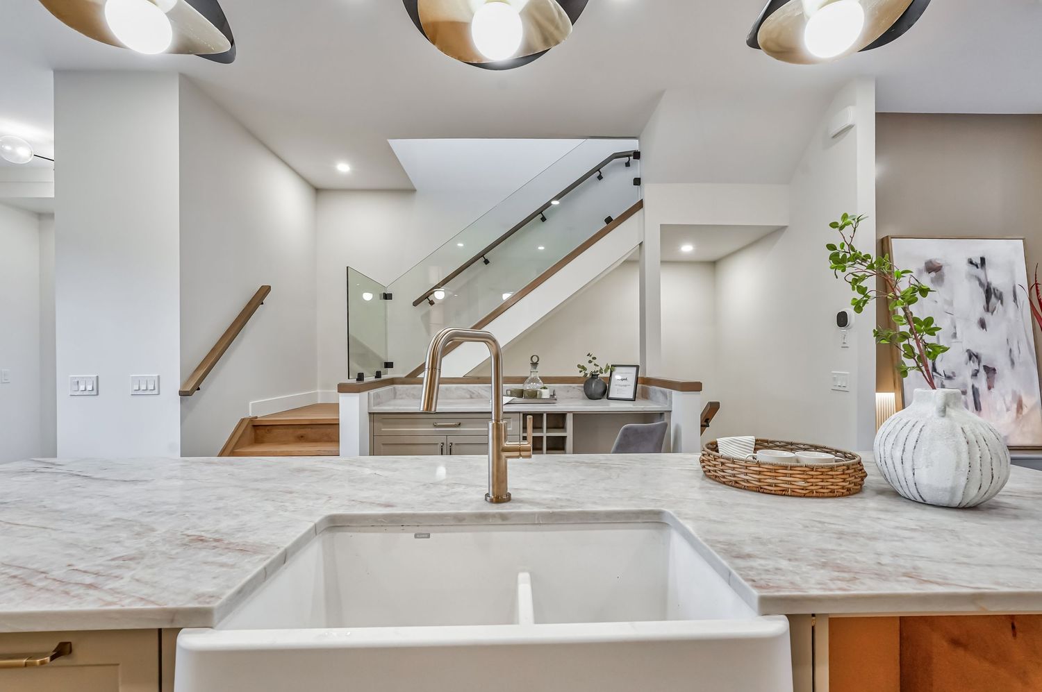 Modern kitchen featuring a farmhouse sink, marble countertop, bronze faucet, and open staircase in a bright white interior.