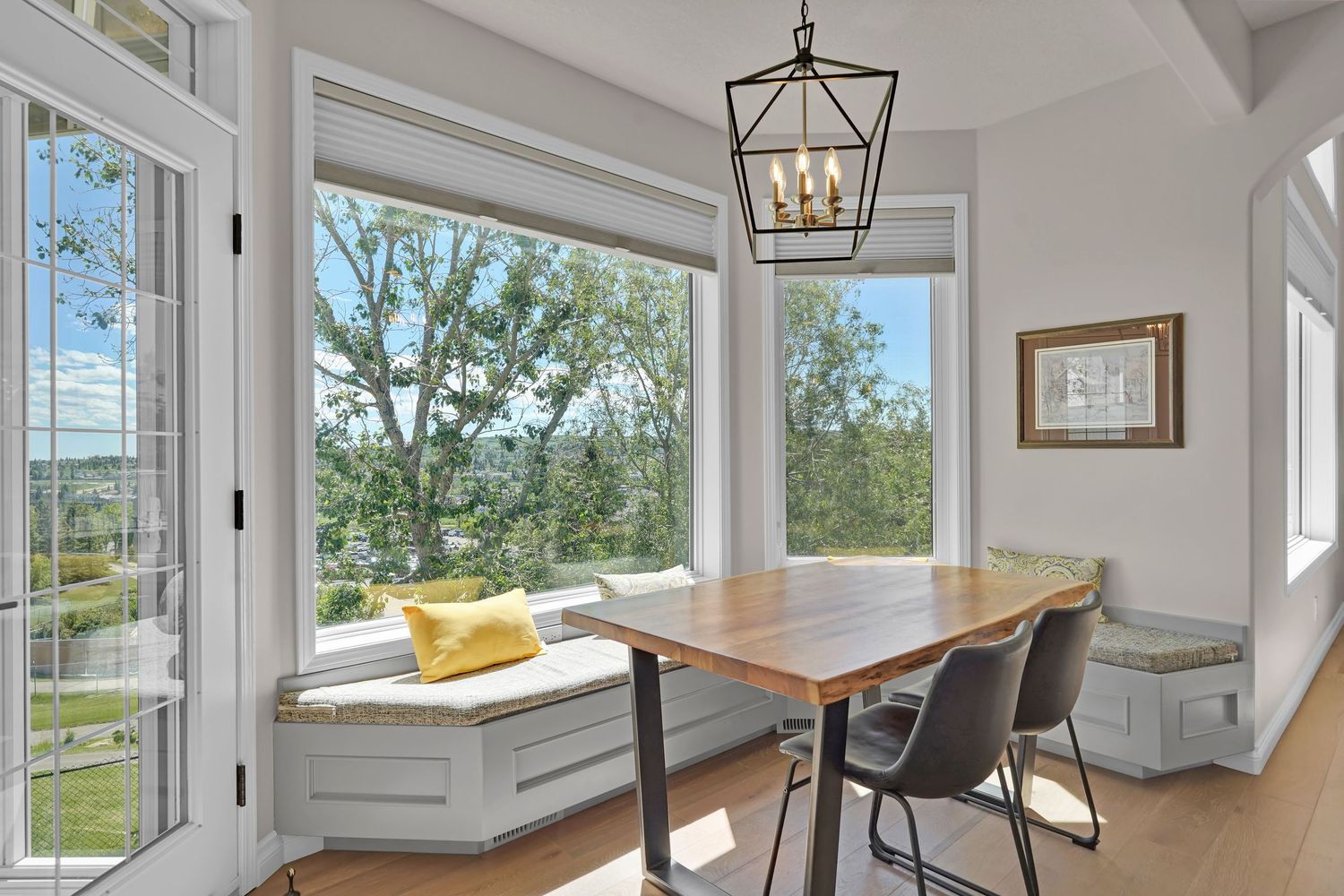 Bright dining nook with wood table, modern chairs, built-in window seat, and large windows overlooking lush green trees.