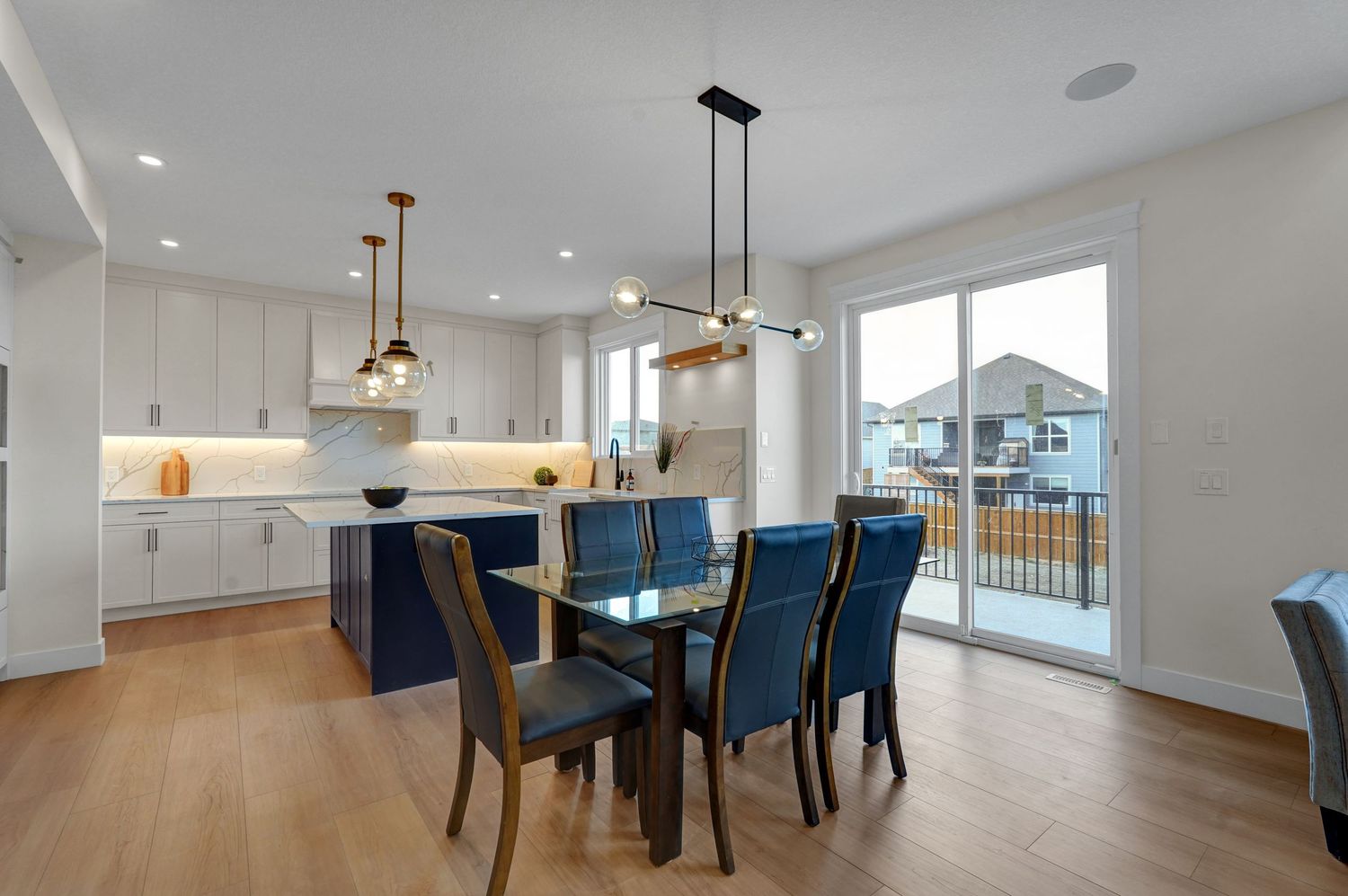 Open dining area with glass table, blue chairs, pendant lights, white kitchen, and patio door access.