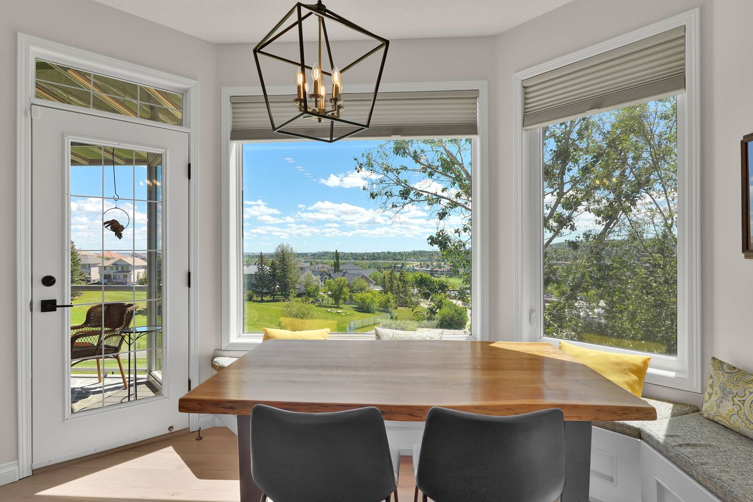 Bright dining nook with wood table, modern chandelier, and panoramic windows overlooking a lush green landscape.