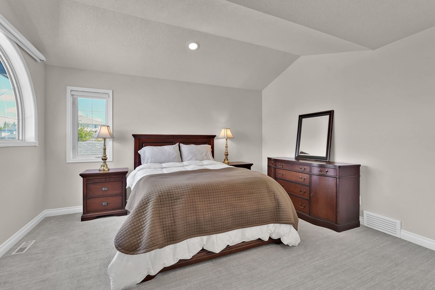 Spacious master bedroom with vaulted ceiling, dark wood furniture, neutral gray walls, and plush carpet flooring.