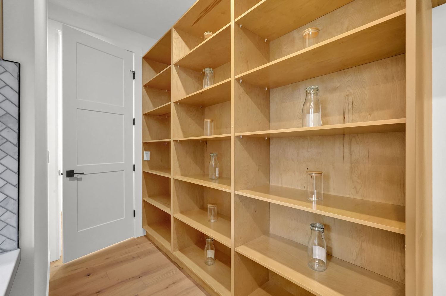 Built-in natural wood pantry shelving unit with multiple open shelves and glass bottles for storage.