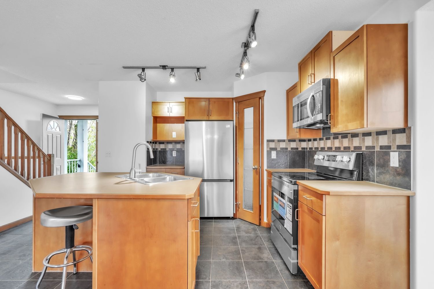 Kitchen with warm wood cabinets, island with sink, stainless steel appliances, and dark tile floors.