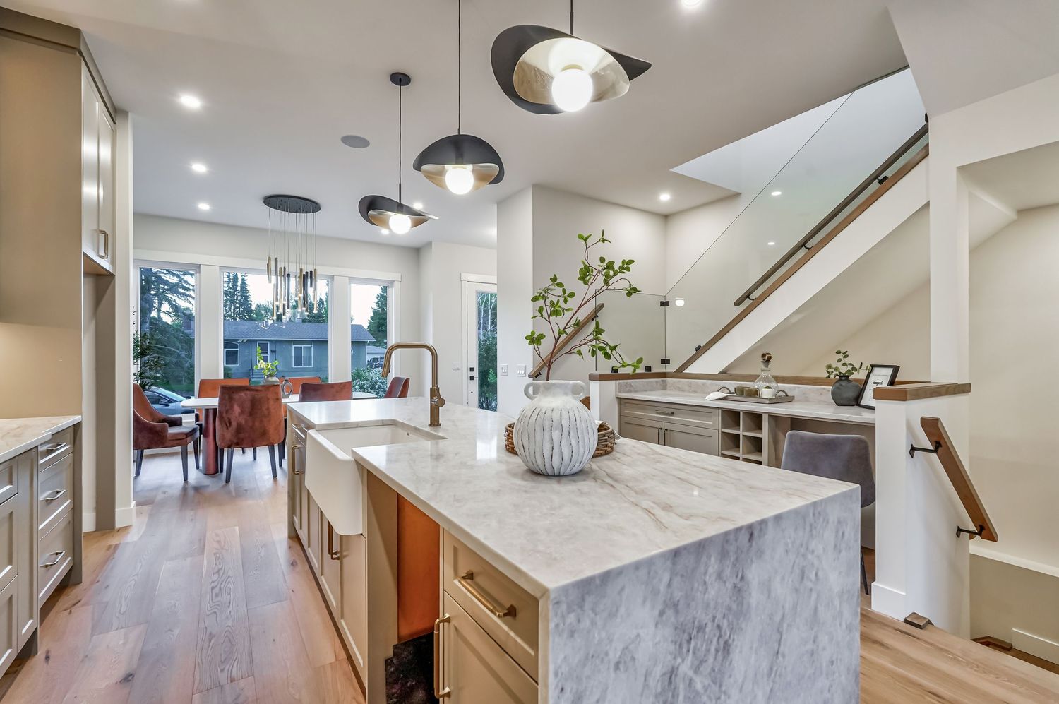 Modern open-concept kitchen with marble island, pendant lights, hardwood floors, and staircase in a bright luxury home.