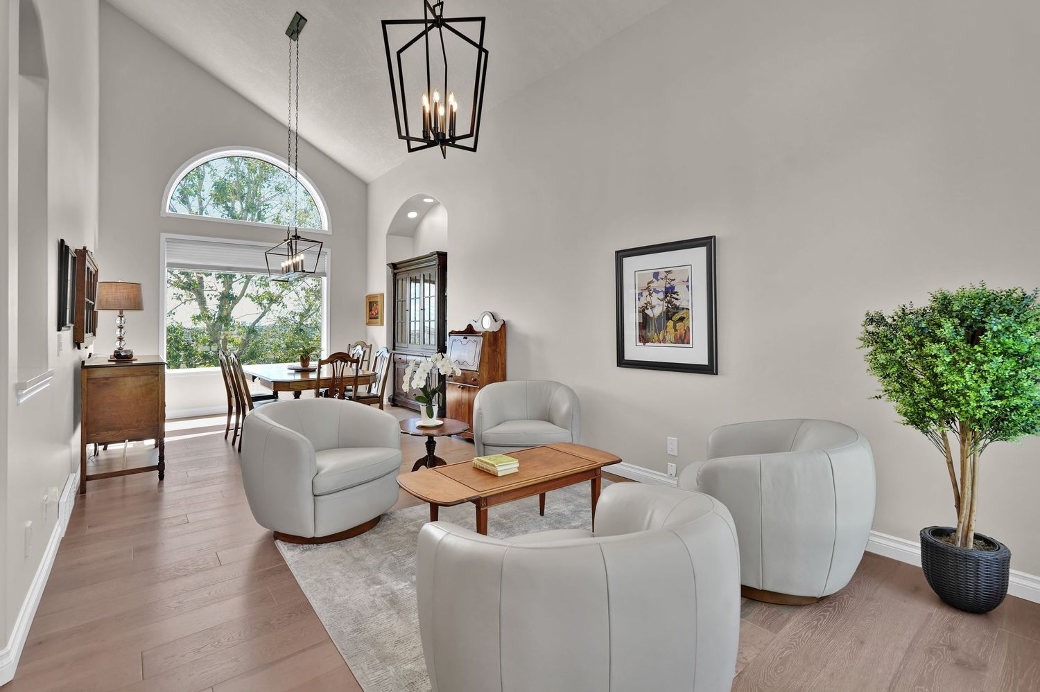 Bright living room with vaulted ceilings, gray curved chairs, wood coffee table, arched window, chandelier, and potted tree.