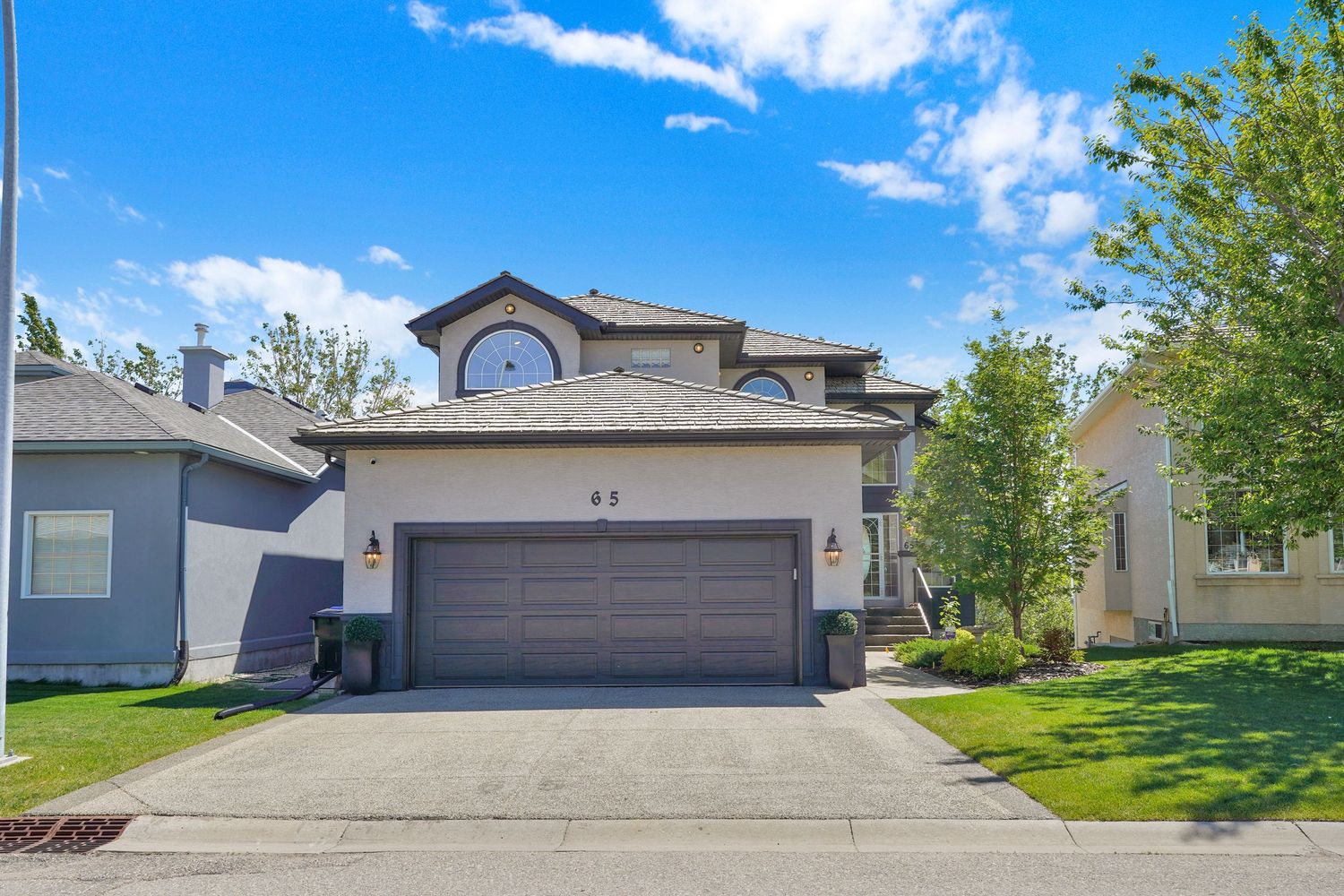 Two-story suburban home with brown garage door, circular window, manicured lawn, and tree-lined street on a sunny day.