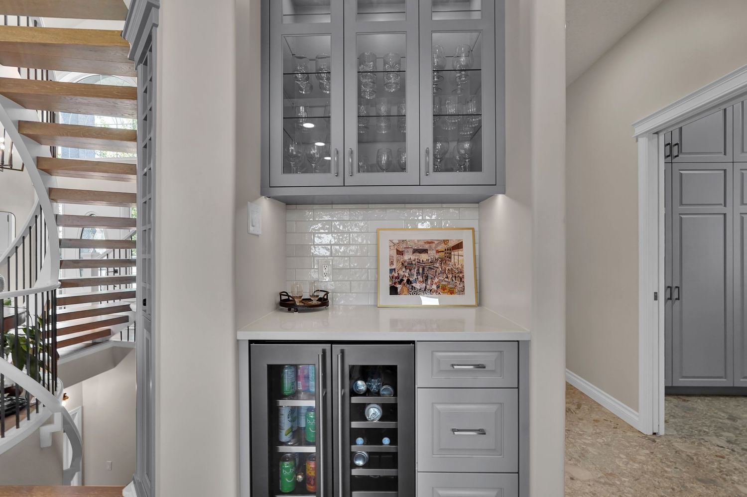 Gray built-in bar cabinet with glass doors, wine fridge, and white subway tile backsplash near a staircase.