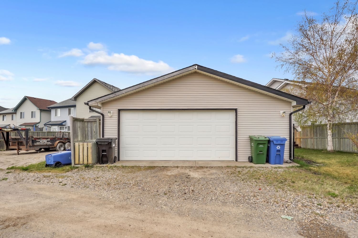 Detached double-car garage with white door, gravel driveway, and green bins beside a wood fence.